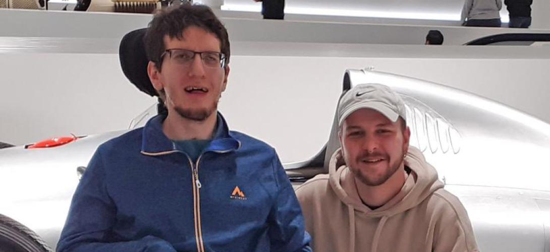 Auf dem Bild sind zwei lächelnde junge Männer vor einem silbernen Sportwagen zu sehen. Einer sitzt im Rollstuhl, der andere kniet neben ihm.