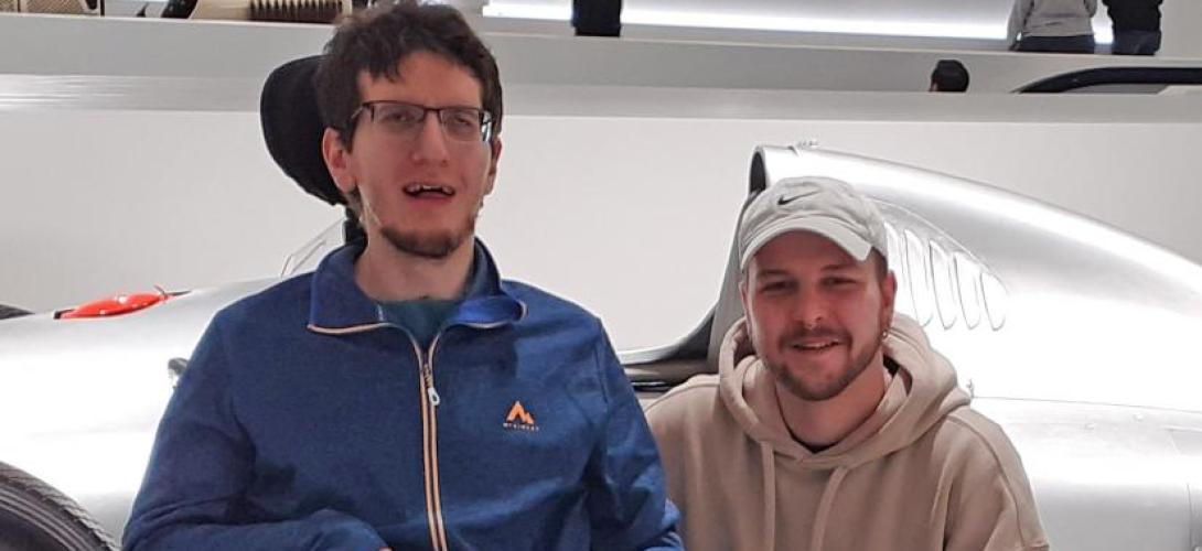Auf dem Bild sind zwei lächelnde junge Männer vor einem silbernen Sportwagen zu sehen. Einer sitzt im Rollstuhl, der andere kniet neben ihm.