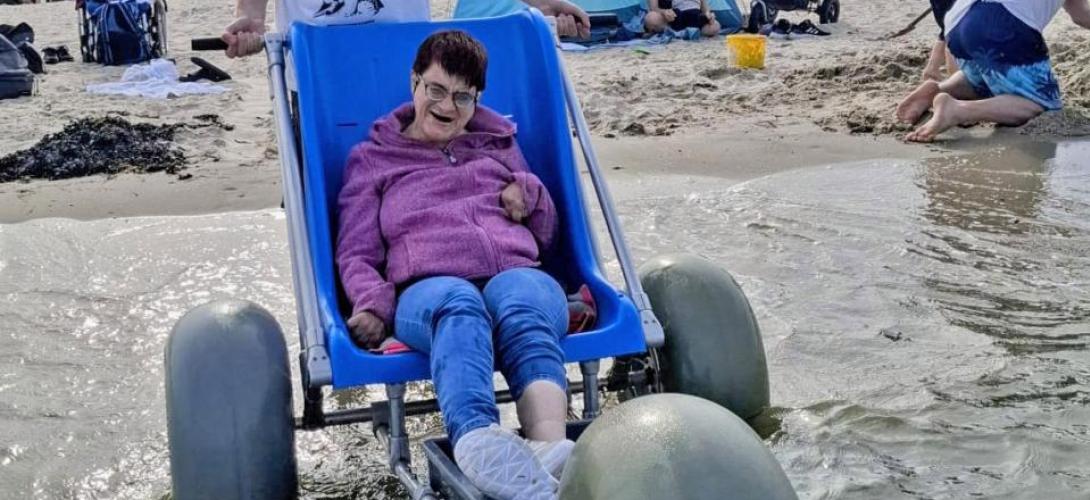 Ein Bild am Nordseestrand in Holland. Eine Frau sitzt am Meer an der Wasserkante im Strandrolli mit großen Ballonreifen. Sie lacht in die Kamera.