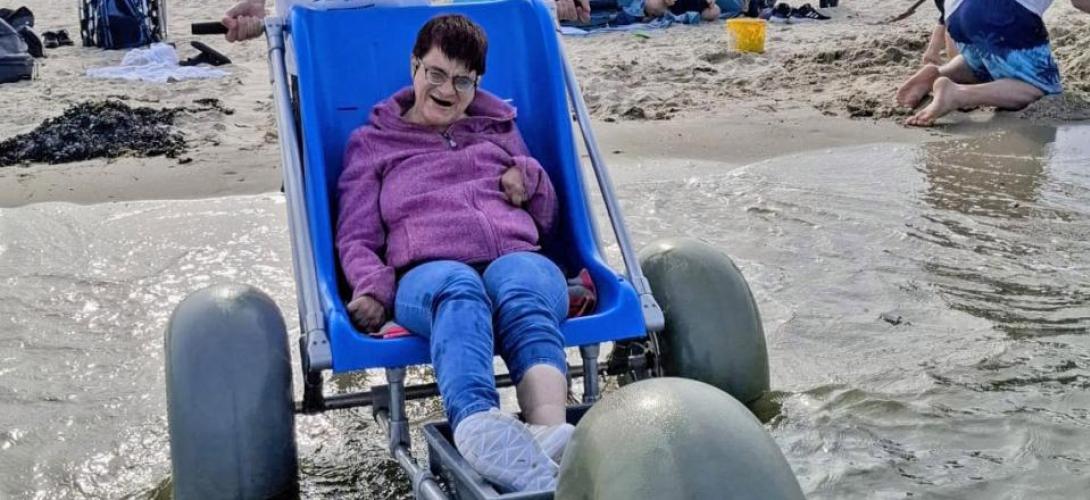 Ein Bild am Nordseestrand in Holland. Eine Frau sitzt am Meer an der Wasserkante im Strandrolli mit großen Ballonreifen. Sie lacht in die Kamera.
