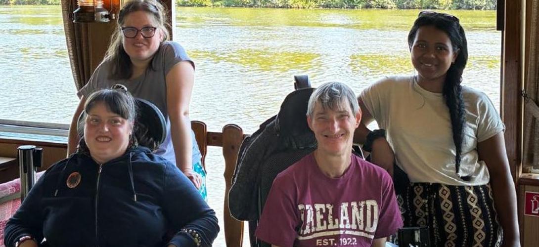 Zu sehen sind vier Frauen, zwei davon im Rollstuhl. Sie lachen in die Kamera. Im Hintergrund fließt ein Fluss.