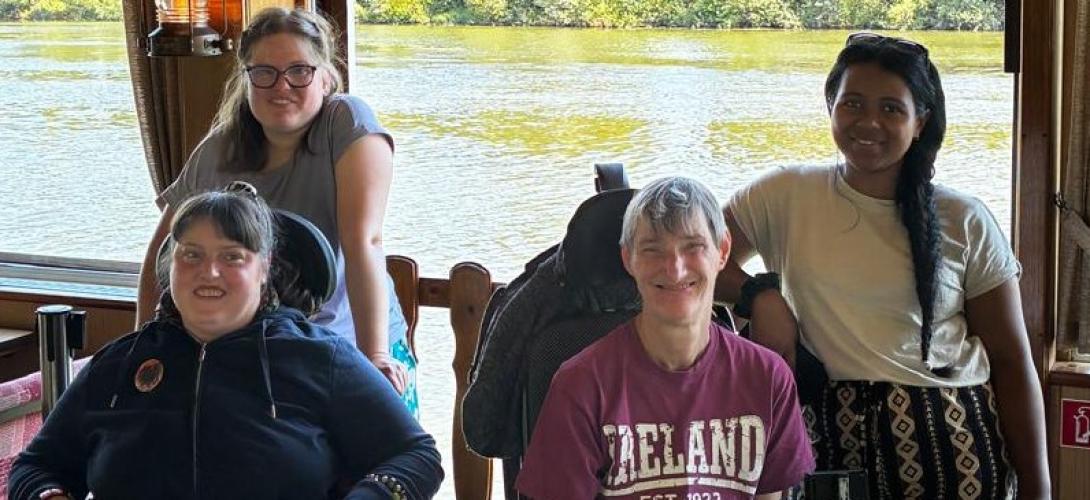 Zu sehen sind vier Frauen, zwei davon im Rollstuhl. Sie lachen in die Kamera. Im Hintergrund fließt ein Fluss.