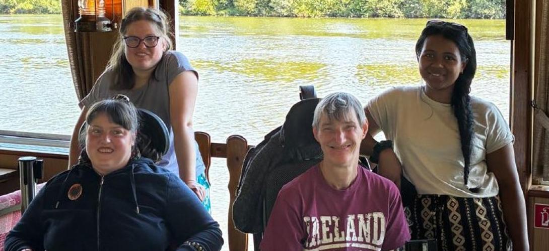 Zu sehen sind vier Frauen, zwei davon im Rollstuhl. Sie lachen in die Kamera. Im Hintergrund fließt ein Fluss.