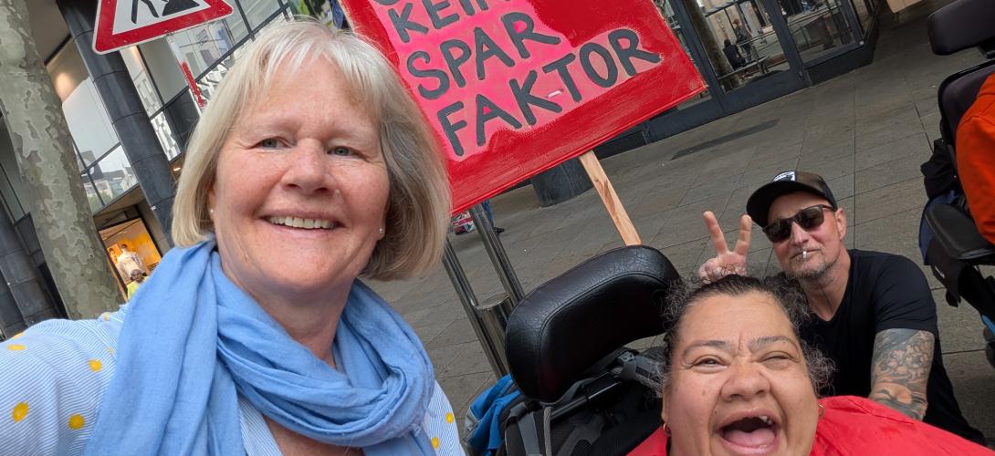 Auf dem Bild demonstrieren Menschen mit und ohne Rollstuhl gegen die geplanten Sparmaßnahmen der Bundesregierung. Sie halten ein rotes Schild hoch. Darauf steht: "Wir sind kein Sparfaktor"
